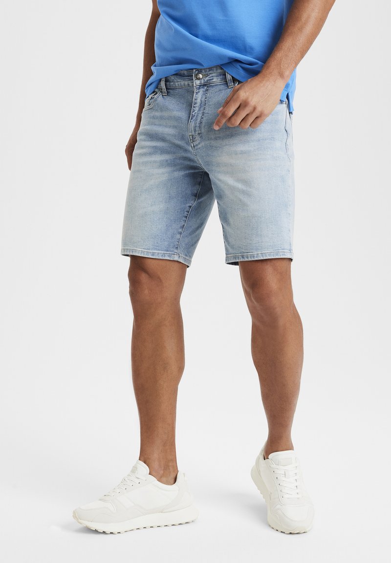Light blue denim shorts with a straight cut, five pockets, and visible stitching, paired with white sneakers and a blue t-shirt.