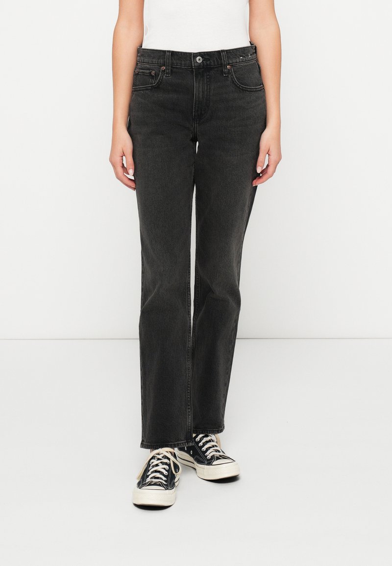 Jean en denim noir taille haute avec une coupe droite, cinq poches et des détails de couture subtils, portés avec des baskets noires et blanches.