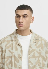Chemise beige à boutons avec des motifs abstraits beiges, dotée d'une poche sur la poitrine et d'un col, portée par-dessus un t-shirt blanc à col rond.