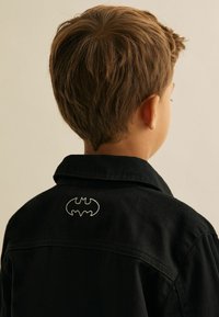 Mustan denimi jope, millel on krae, ning seljal on valge tikandiga Batmani logo. Tekstuur on sile, nähtavad õmbluse üksikasjad.