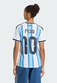 Žena s kudrnatými vlasy v drdolu, která má na sobě argentinský fotbalový dres s nápisem "MESSI" a číslem 10 na zádech, spojený s modrými džínami.