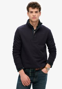 Jonge man die een marineblauwe quarter-zip pullover over een groen geruit shirt draagt, blauwe jeans en een bruine leren horloge om zijn linkerpols.