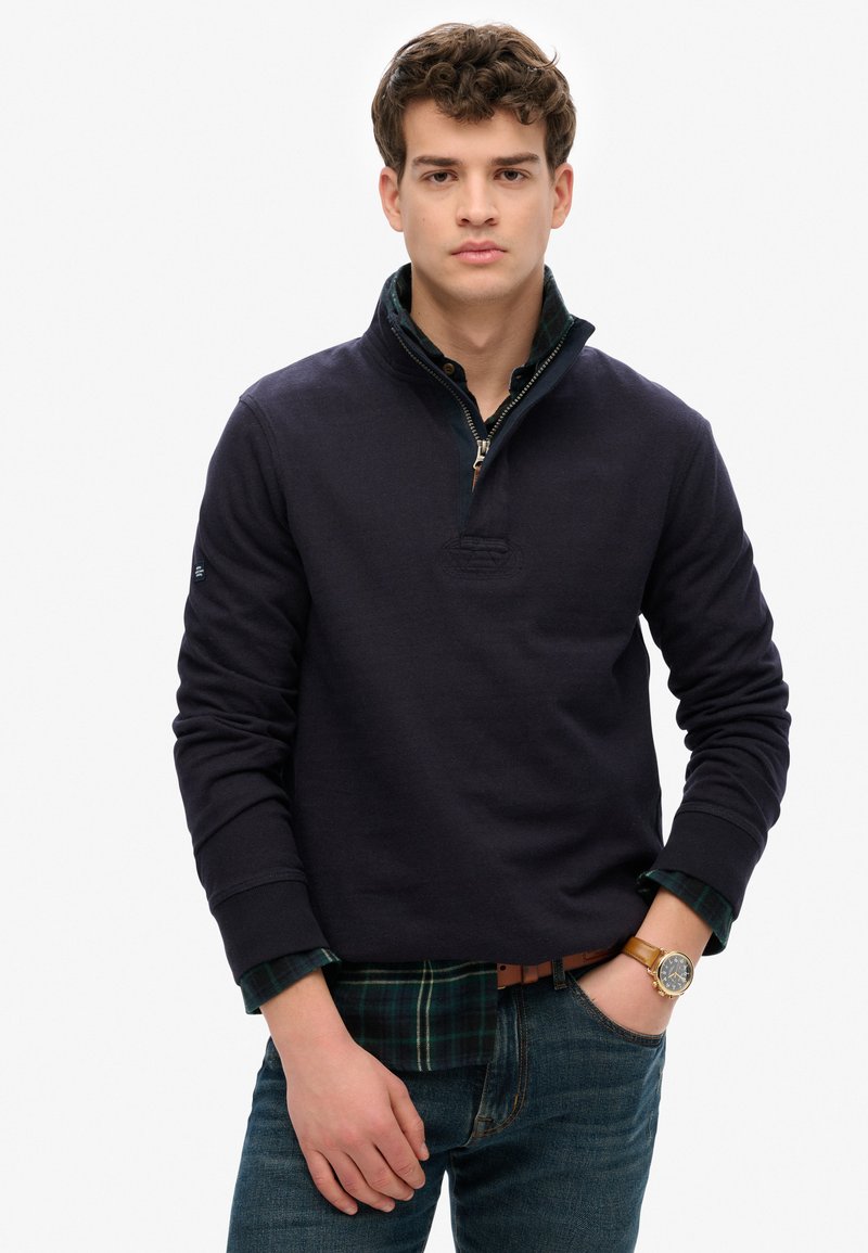 Jonge man die een marineblauwe quarter-zip pullover over een groen geruit shirt draagt, blauwe jeans en een bruine leren horloge om zijn linkerpols.