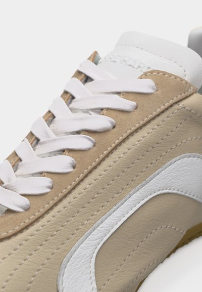 Gros plan sur une basket beige et blanche avec des lacets blancs, une texture en cuir et des coutures détaillées sur des surfaces lisses.