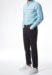 Hombre con camisa de cuadros azul claro, pantalones negros arremangados, zapatillas blancas, de pie con las manos en los bolsillos frente a un fondo blanco.