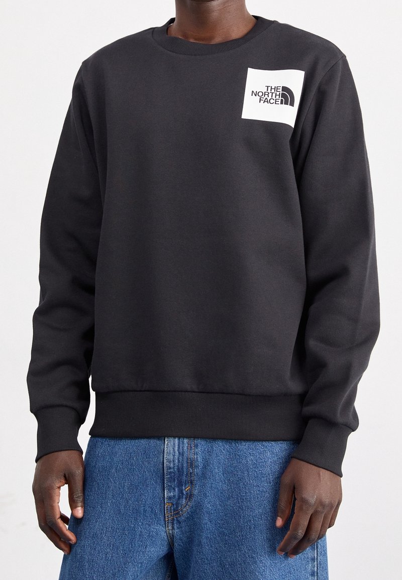 Sweat-shirt noir à col rond avec un logo carré blanc indiquant "The North Face" porté avec un jean bleu sur une personne à la peau foncée.