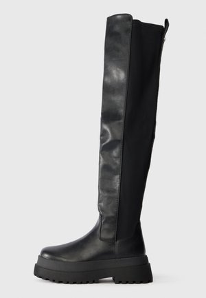 Black knee-high boot with chunky sole, smooth leather front, elastic back panel, and pull tab at the top on a plain background.