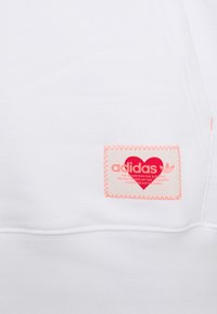 White sweatshirt with a textured fabric featuring a prominent pink Adidas label stitched on, displaying a heart and three stripes logo.