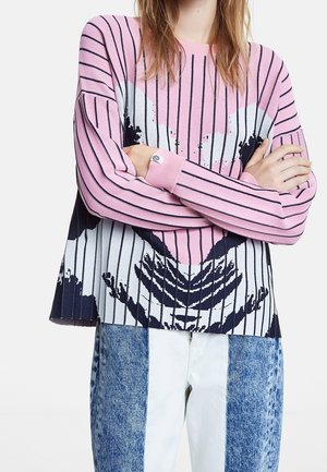 Persona con suéter de rayas rosas, blancas y negras con diseño de corazón, combinado con vaqueros patchwork azules y blancos, brazos cruzados sobre el pecho.