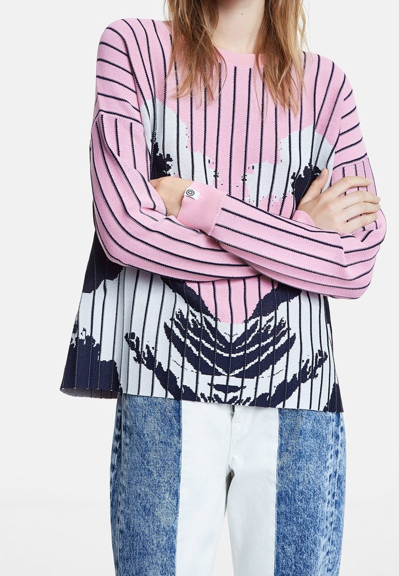 Personne portant un pull rayé rose, blanc et noir avec un motif cœur, associé à un jean patchwork bleu et blanc, les bras croisés sur la poitrine.