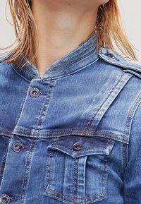 Ljust blå denimjacka med mandarynkrage, med synliga sömmar, knappstängningar och två bröstfickor med klappar.