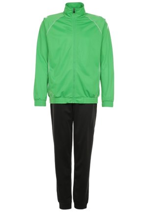 Mężczyzna w jaskrawozielonej kurtce na zamek, zielonej koszulce, czarnych spodniach dresowych i białych trampkach, idący z jedną ręką w kieszeni na szarym tle.