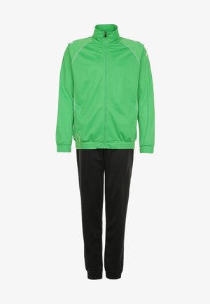 Uomo che indossa una giacca verde acceso con cerniera, maglietta verde, pantaloni da ginnastica neri e scarpe da ginnastica bianche, camminando con una mano in tasca su sfondo grigio.