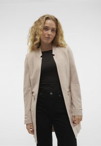 Abrigo largo de color beige claro, ajustado, con textura de espiga, frente abierto y bolsillos con cremallera, combinado con una blusa negra y pantalones oscuros.