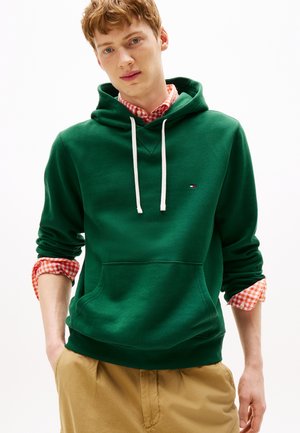 Jeune homme portant un sweat à capuche vert avec des cordons blancs, une chemise à carreaux orange avec les manches retroussées et un pantalon beige, debout les mains dans les poches.