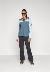 Lichtblauwe en donkerblauwe zippered jas met capuchon, gecombineerd met zwarte broeken; beide gemaakt van duurzame materialen. Bruine wandelschoenen.