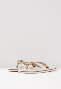 Tan flip-flops met gekruiste bandjes en gouden hardware-details, met een gestructureerde witte zool en contrasterende bruine randen.