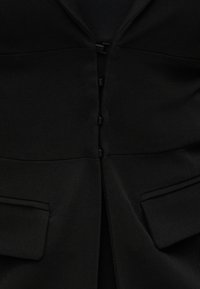 Blazer noir avec un col cranté, fermeture avant à cinq boutons noirs et deux poches inclinées. Tissu à texture lisse.