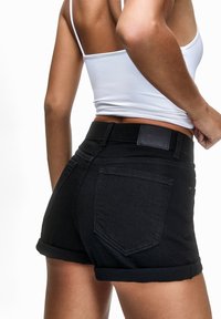 Musta farkkushortsit käärityillä lahkeilla, joissa on kaksi takataskua ja nahkainen etiketti vyötäröllä. Minimalistinen muotoilu puhtain linjoin.