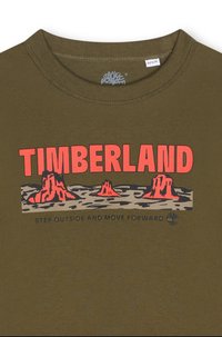 Olijfgroene katoenen T-shirt met een groot rood "TIMBERLAND" logo, vergezeld van een afbeelding van bergen en de tekst "GA BUITEN EN GA VOORUIT."