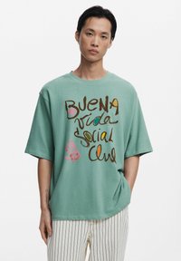 Mint green oversized t-shirt with "BUENA VIDA SOCIAL CLUB" graphic in multicolored text. Short sleeves and relaxed fit, worn with striped pants.