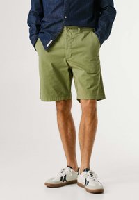 Άνδρας που φοράει χακί σορτς, τζιν πουκάμισο με τα χέρια στις τσέπες και λευκά αθλητικά παπούτσια με σκούρες μπλε λεπτομέρειες, στέκεται σε απλό φόντο.