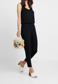 Mono negro sin mangas con cintura ceñida, combinado con sandalias blancas de tiras tejidas y una bolsa redonda de mimbre con bordado floral.
