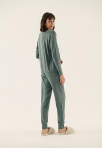 Teal Loungewear-Set aus weichem Stoff; mit langen Ärmeln, locker sitzendem Oberteil, schmal zulaufenden Hosen mit gerippten Bündchen, kombiniert mit beigen Hausschuhen.