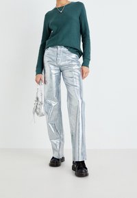 Personne portant un haut à manches longues turquoise, un pantalon argenté métallisé, des bottes noires, tenant un petit sac à main argenté sur un fond blanc uni.