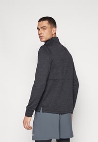 Haut de sport à manches longues gris foncé avec col montant, présentant un design épuré et une texture contrastante à l'arrière, associé à un short gris clair.