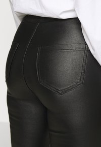Svart imitationläderbyxor med hög midja, med två bakfickor och subtila söm-detaljer. Slät textur med en glansig yta.
