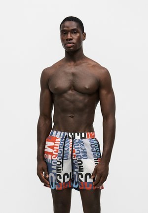 Homme musclé torse nu portant un short de bain multicolore avec un grand texte de la marque Moschino, debout sur un fond blanc uni.