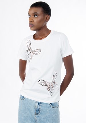 Giovane donna che indossa una T-shirt bianca con ritagli di pizzo a forma di farfalla e jeans azzurri, che guarda verso sinistra con le mani dietro la schiena.