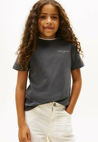 Pige med langt, krøllet hår iført en mørkegrå Tommy Hilfiger t-shirt og hvide bukser, stående med den ene hånd i lommen.