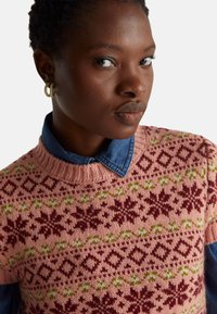 Pletený svetřík světle růžové barvy s vzorovaným motivem v bordó a zelené. Nositelný přes džínovou košili s límcem.