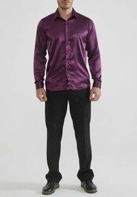 Chemise violette brillante à manches longues avec un col boutonné, des boutons sur le devant et une texture lisse ; associée à un pantalon et des chaussures noires.