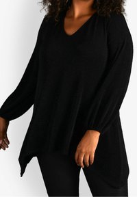 Túnica negra con cuello en V hecha de una tela texturizada con un brillo sutil. Mangas largas y abullonadas. Dobladillo asimétrico con un corte relajado.