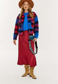 Kolorowa kurtka w kratę w odcieniach czerwieni, niebieskiego i czarnego; niebieski sweter; czerwona maxi spódnica; beżowe kozaki za kolano; dekoracyjny pasek torby.