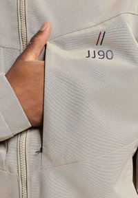 Lys beige jakke med teksturert stoff, med glidelås og en lomme som viser "JJ90" i mørkegrå og røde detaljer.