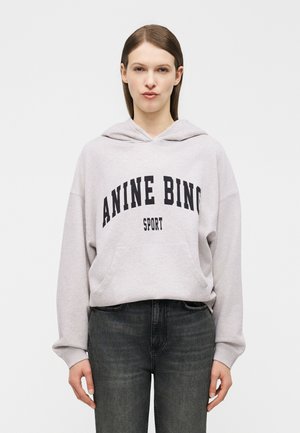 Woman with straight brown hair wearing a light gray hoodie with "ANINE BING SPORT" text and dark gray jeans against a plain background.