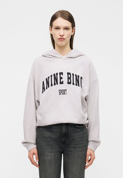 Mujer con cabello castaño lacio vistiendo una sudadera con capucha gris claro con el texto "ANINE BING SPORT" y jeans gris oscuro, sobre un fondo liso.