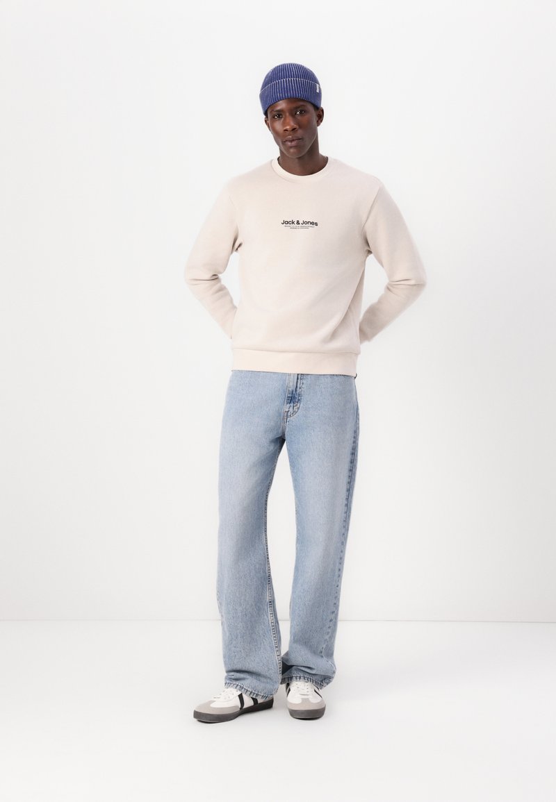 Beige sweatshirt met een zwart logo, lichtblauwe wijde jeans en grijze sneakers met zwarte strepen. Model draagt een donkerblauwe beanie.