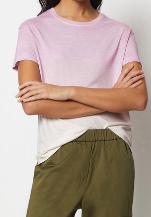 Personne les bras croisés portant un t-shirt à manches courtes dégradé rose à blanc et un pantalon taille haute vert olive.