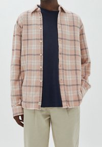 Camisa de franela a cuadros rosa y crema con diseño de botones, llevada sobre una camiseta azul marino, combinada con pantalones de color claro.