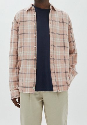Roze en crèmekleurig geruit flanellen overhemd met een knoopsluiting, gedragen over een marineblauw T-shirt, gecombineerd met lichtgekleurde broek.