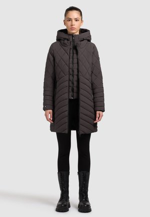 Gevoerde grijze winterjas met een capuchon, voorzien van diagonale patronen en een rits aan de voorkant, gecombineerd met zwarte leggings en kniehoge laarzen.