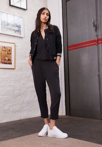 Žena stoji u zatvorenom prostoru noseći crnu bomber jaknu, crne hlače, crni top i bijele tenisice, s uokvirenim fotografijama na bijelom zidnom zidu od opeke.