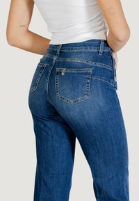 Jeans di denim blu a vita alta con gamba dritta, cinque tasche, cuciture sottili e dettaglio con bottone argento in vita.