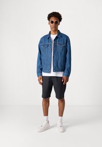 Denim jacket in blue, featuring two chest pockets, buttons, and a patterned texture. Worn over a white t-shirt, paired with dark shorts and sneakers.