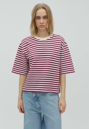 Jeune femme aux cheveux blonds mi-longs portant un t-shirt à manches courtes rayé blanc et bordeaux et un jean bleu clair, debout devant un fond uni.